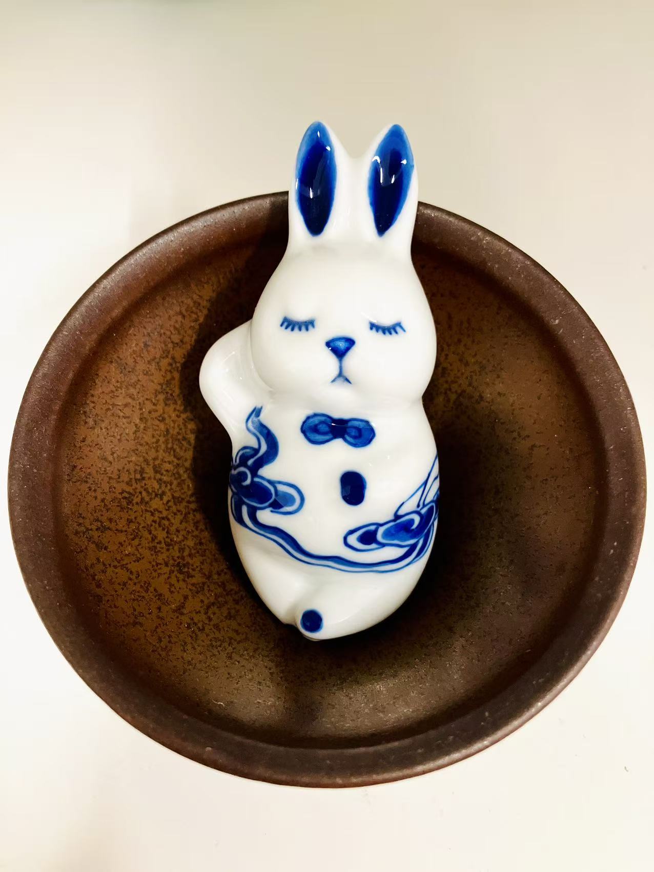 Closed-eye rabbit with blue ears, photographed in its original brown bowl setting